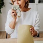 Woman joyfully holding cash and envelope at home desk, savoring her earnings.